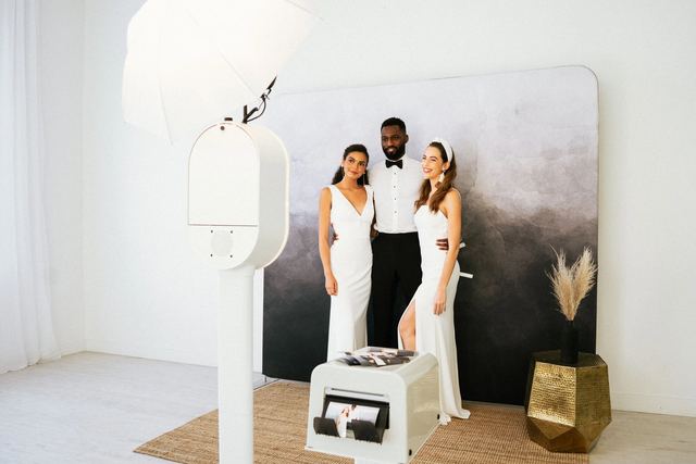 Three guests in formal white and black attire posing beside a white photo booth and print station in front of a gray backdrop