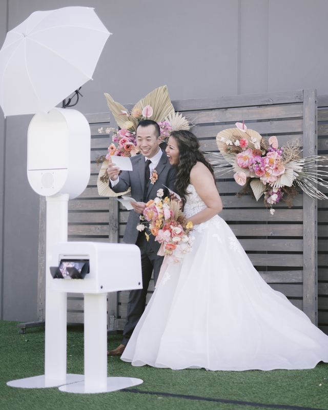 Bride and groom laughing beside a white photo booth in front of a floral backdrop